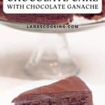 A slice of flourless chocolate cake with chocolate ganache on a plate, whole cake in background on a stand.