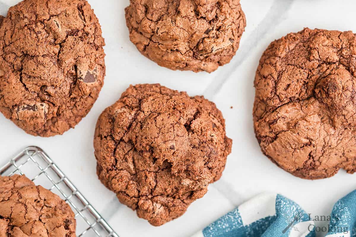 Five chocolate cookies on a white surface, with part of a cooling rack and blue towel visible.