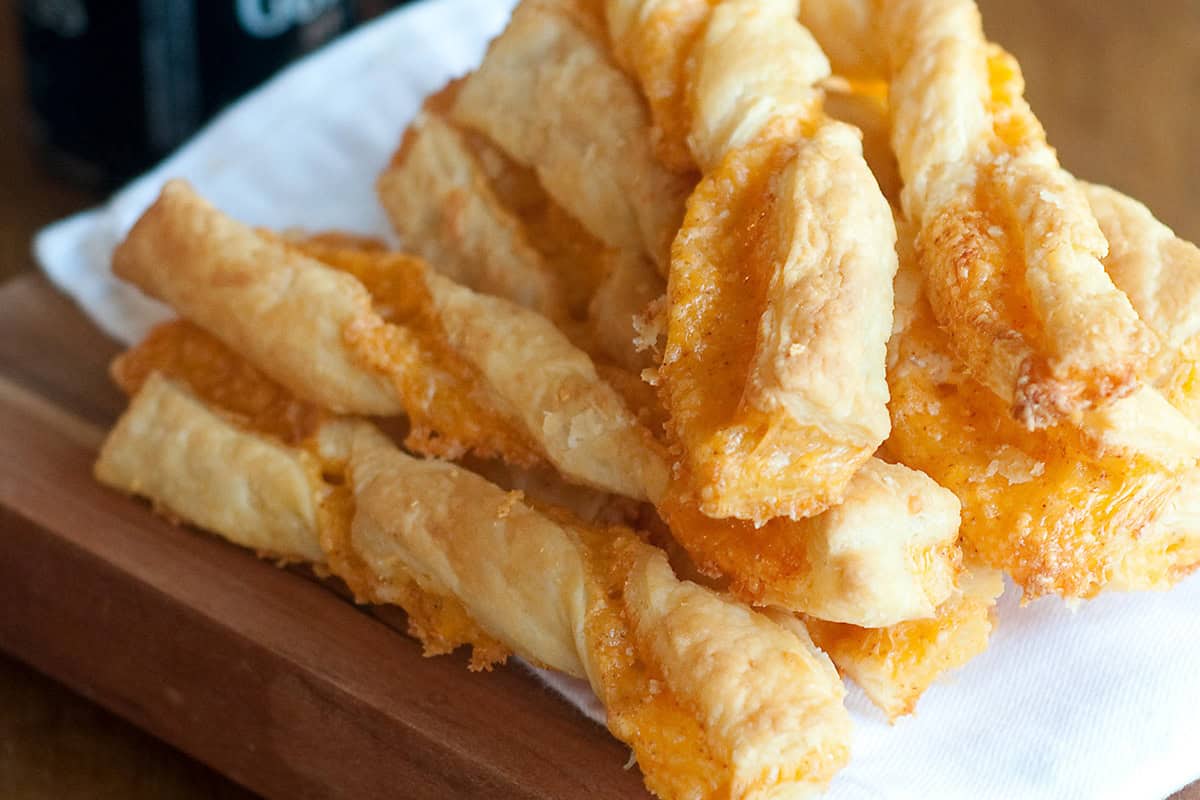 Cheese twists made with puff pastry and melted cheese stacked on a wooden board with a white napkin.