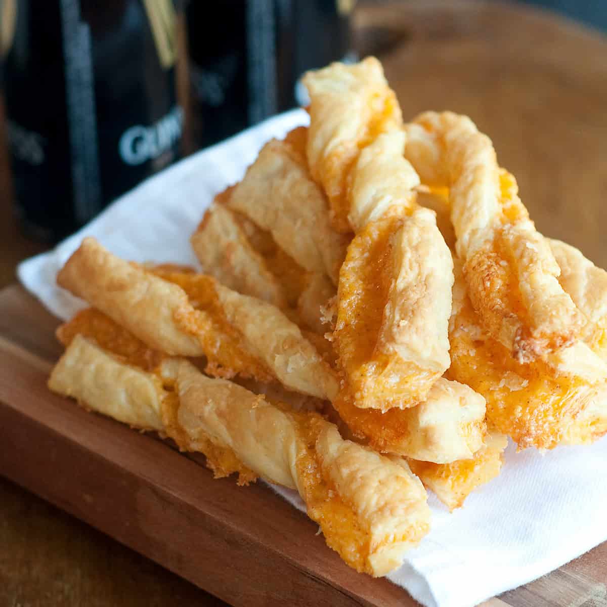 A stack of cheese straws on a white napkin, placed on a wooden board with dark bottles in the background.