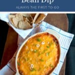 Baked refried bean dip topped with melted cheese and sliced green onions, served with a basket of tortilla chips.