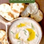 Bowl of white bean dip with olive oil and herbs, surrounded by round crackers on a wooden surface.