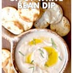 Bowl of white bean dip with olive oil and herbs, served with round crackers on a wooden surface.