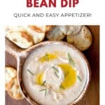 A bowl of spicy white bean dip with olive oil and herbs, served with round crackers on a wooden surface.