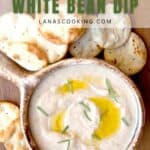 A bowl of white bean dip garnished with olive oil and rosemary, surrounded by round toasted crackers on a wooden surface.