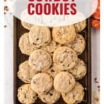 A tray of cowboy cookies with chocolate chips, displayed on a marble surface with a red cloth nearby.