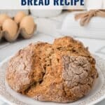 A loaf of brown Irish soda bread on a plate, with eggs and kitchen decor in the background.