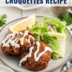 Plate with two salmon croquettes topped with white sauce, salad, and a lemon wedge. Text: Classic Salmon Croquettes Recipe.