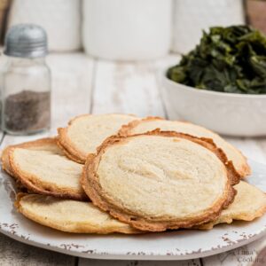 A plate of hoecakes is displayed on a white surface. In the background, there is a white bowl filled with cooked greens and a pepper shaker. The setting has a rustic, home-style appearance.