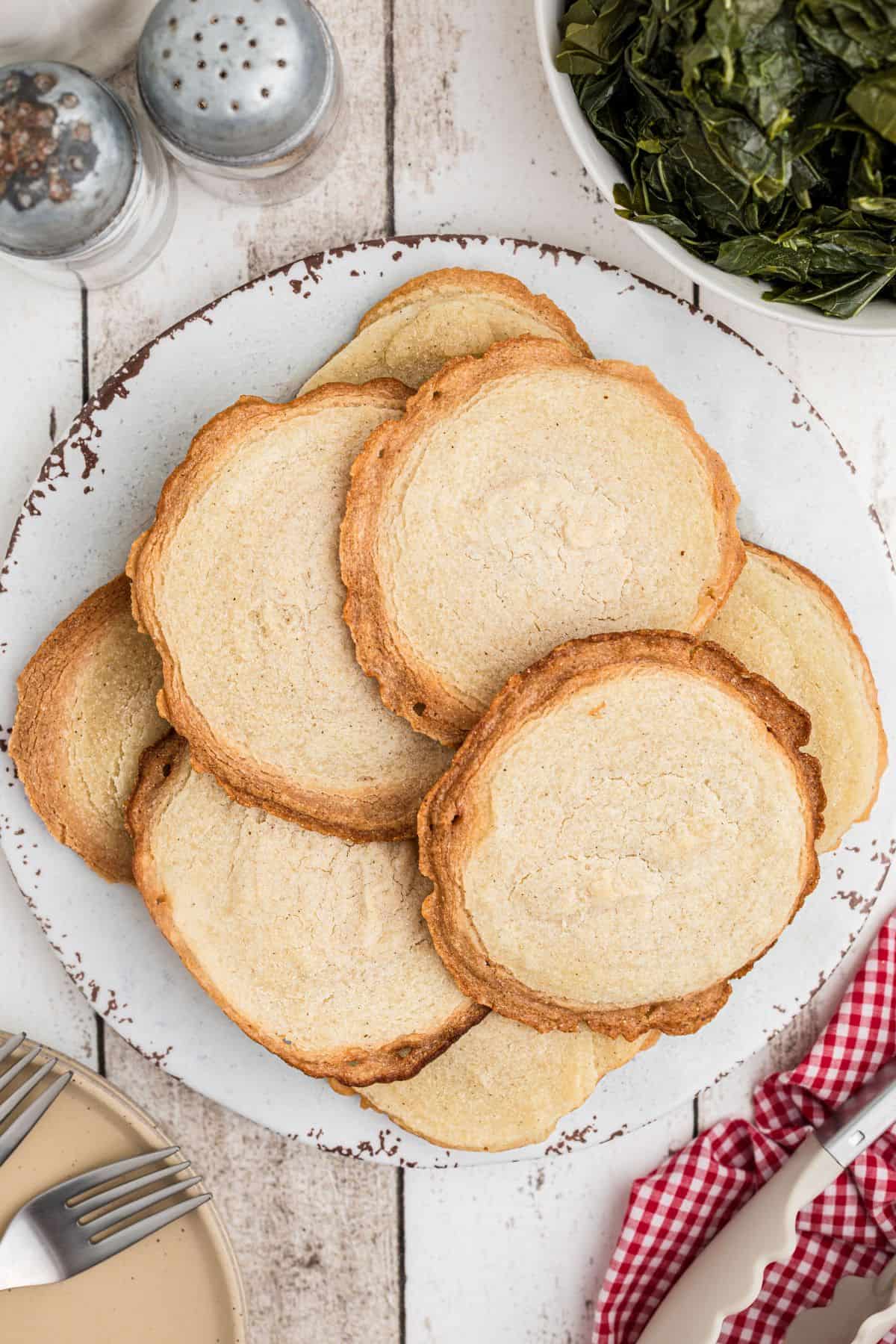 A white plate holds a stack of round, golden brown slices of cornbread. The plate sits on a rustic white wooden table. Collard greens and salt and pepper shakers are nearby. A red and white napkin and plates are partially visible.