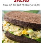 A herbed egg salad sandwich with lettuce on dark bread, shown close up with a fresh, creamy filling.