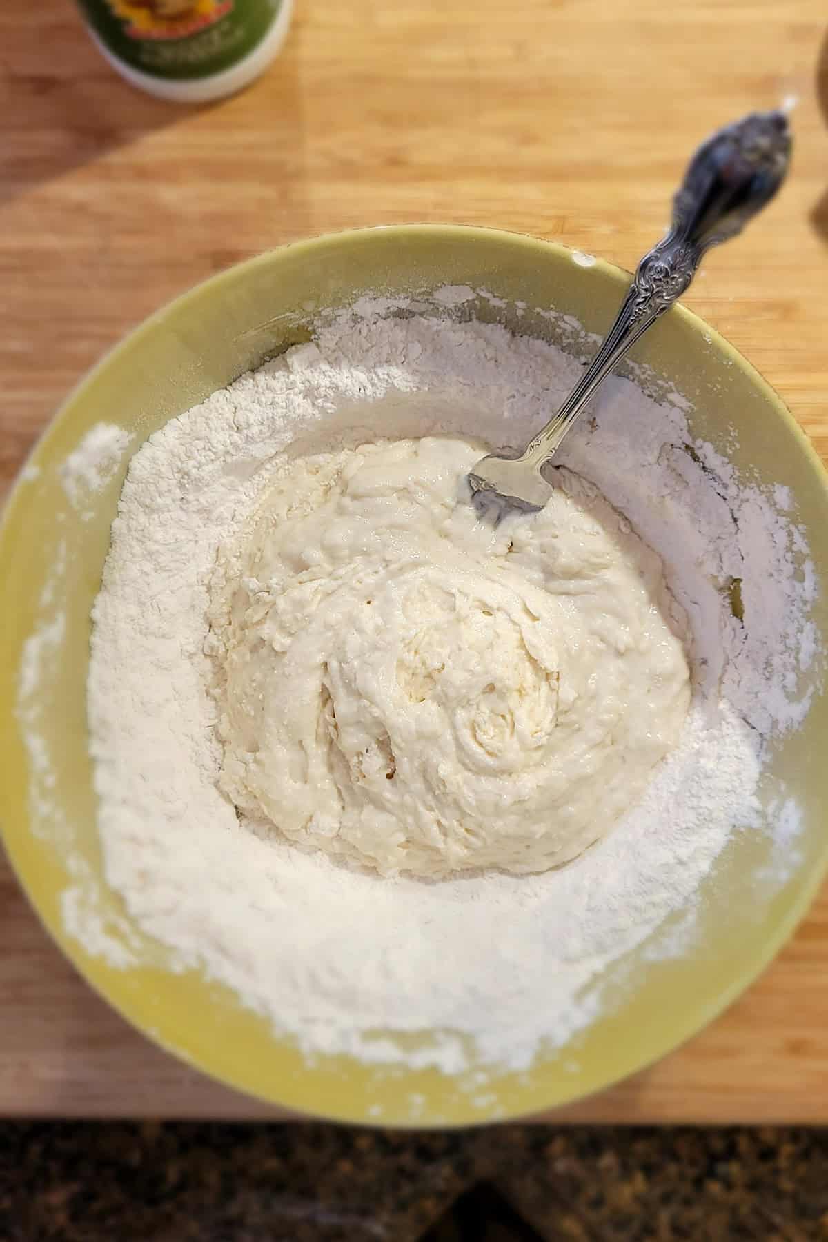 A mixing bowl with flour and wet dough being stirred with a fork on a wooden countertop.