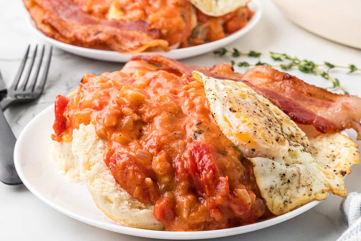 A serving of tomato gravy over biscuits with fried eggs and bacon on the side.