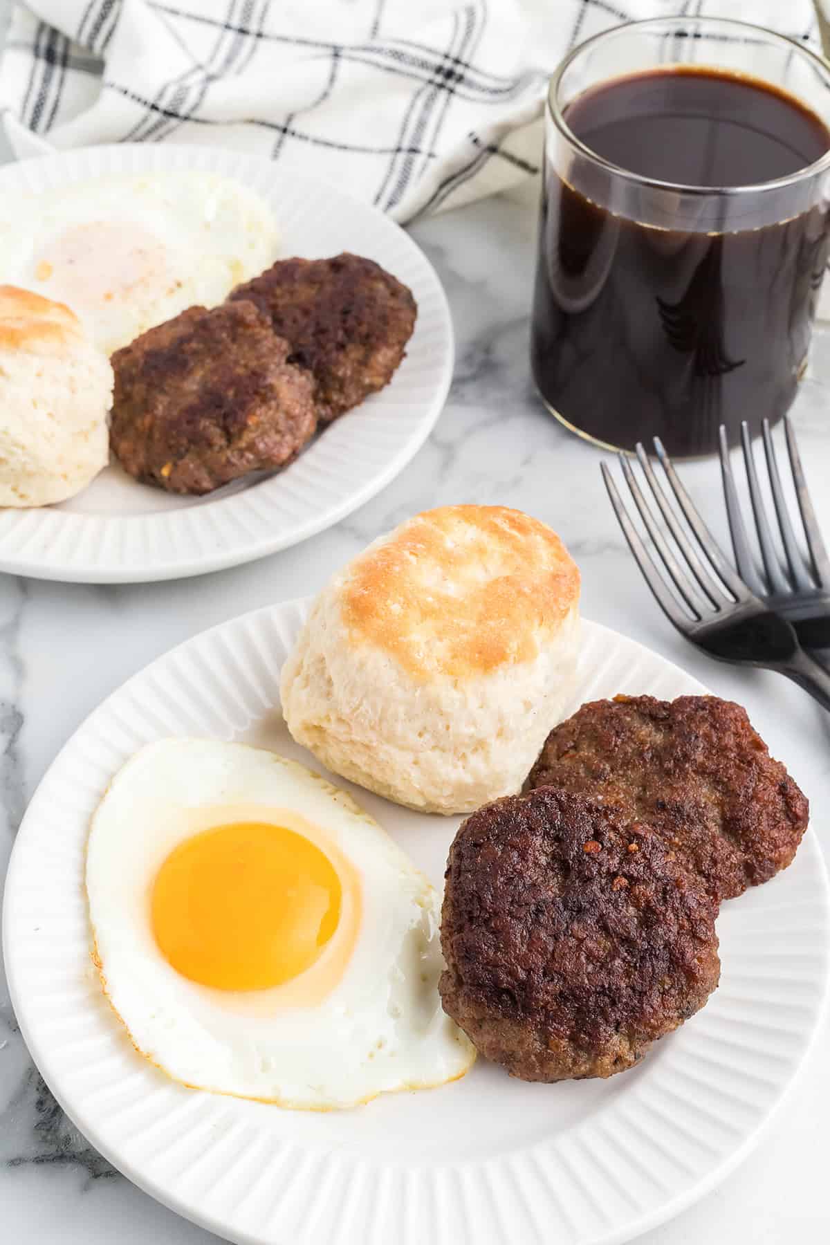 Plate with a sunny-side-up egg, two sausage patties, and a biscuit next to a glass of black coffee.