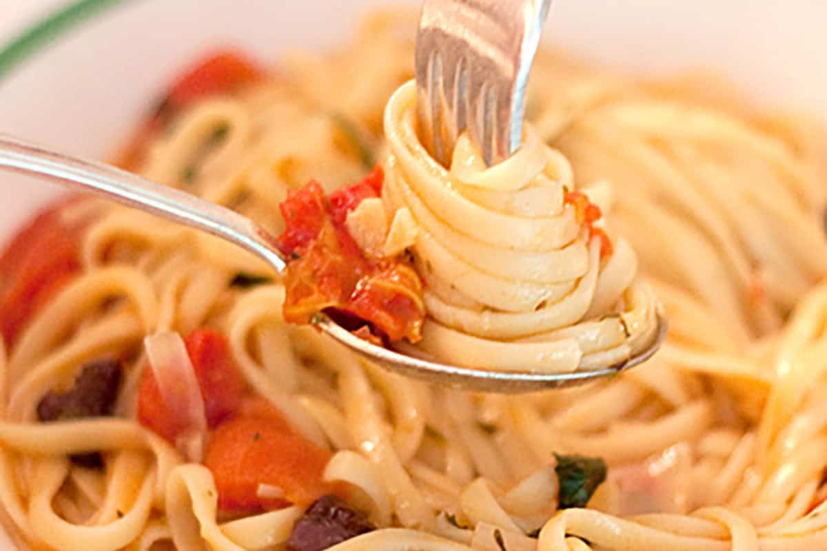 A fork twirls spaghetti with tomato sauce over a spoon, above a bowl of pasta.