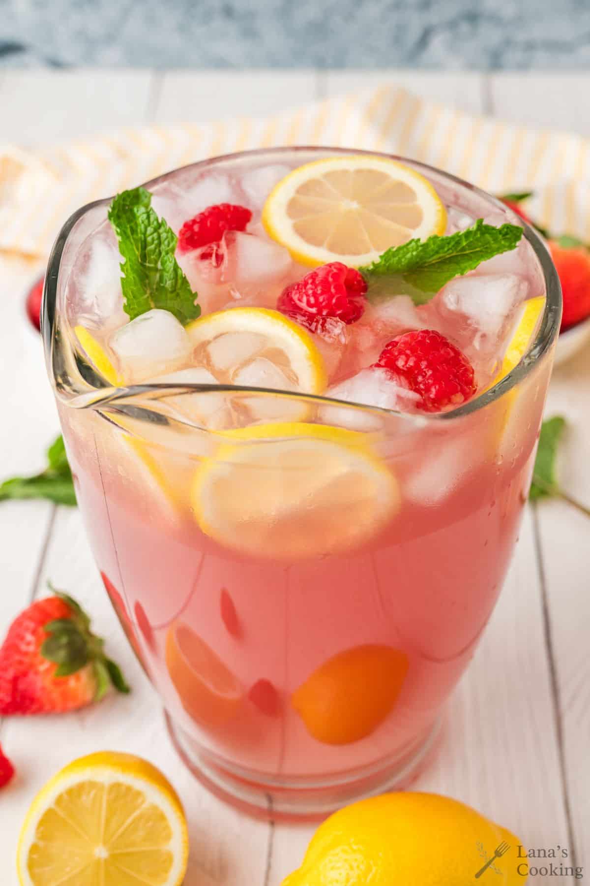 Large glass pitcher of pink lemonade with mint, lemon slices, and strawberries on top.