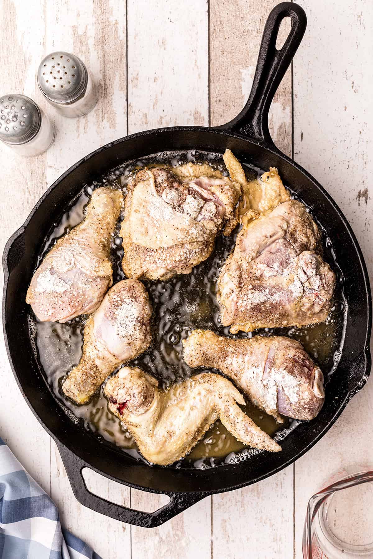 Raw, breaded chicken pieces frying in oil in a black cast iron skillet on a wooden surface.