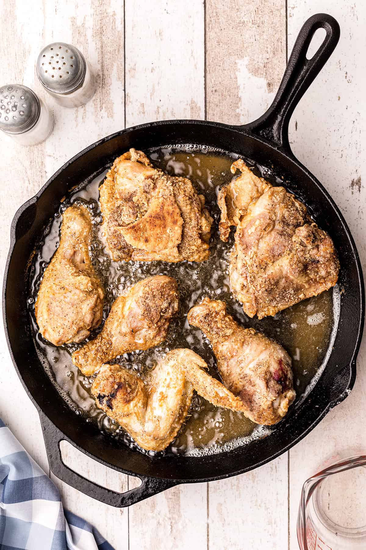Six pieces of fried chicken cooking in a cast iron skillet with oil, next to salt and pepper shakers.