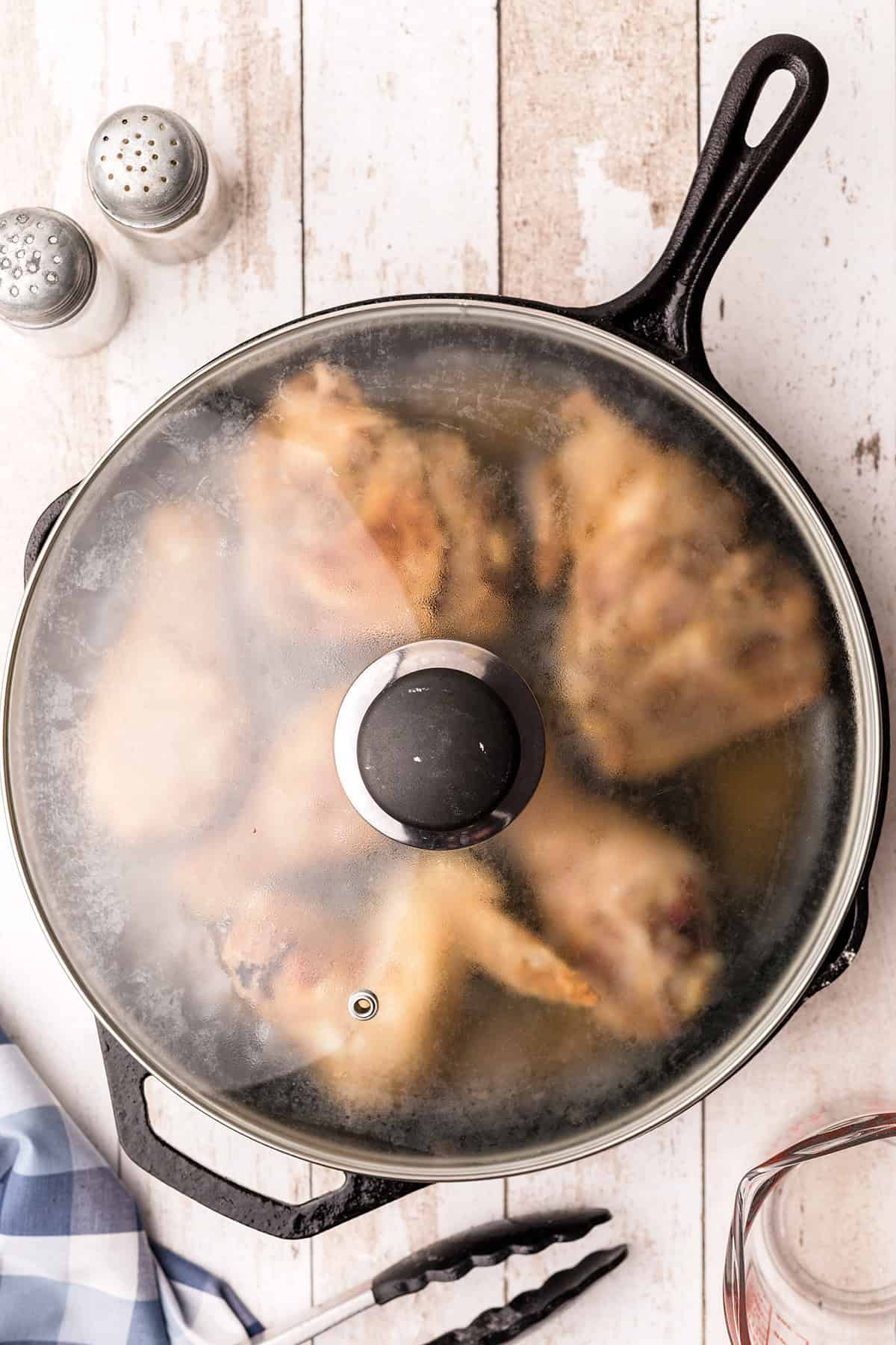 Chicken pieces cooking in a covered skillet on a white surface, with salt, pepper, tongs, and napkin nearby.