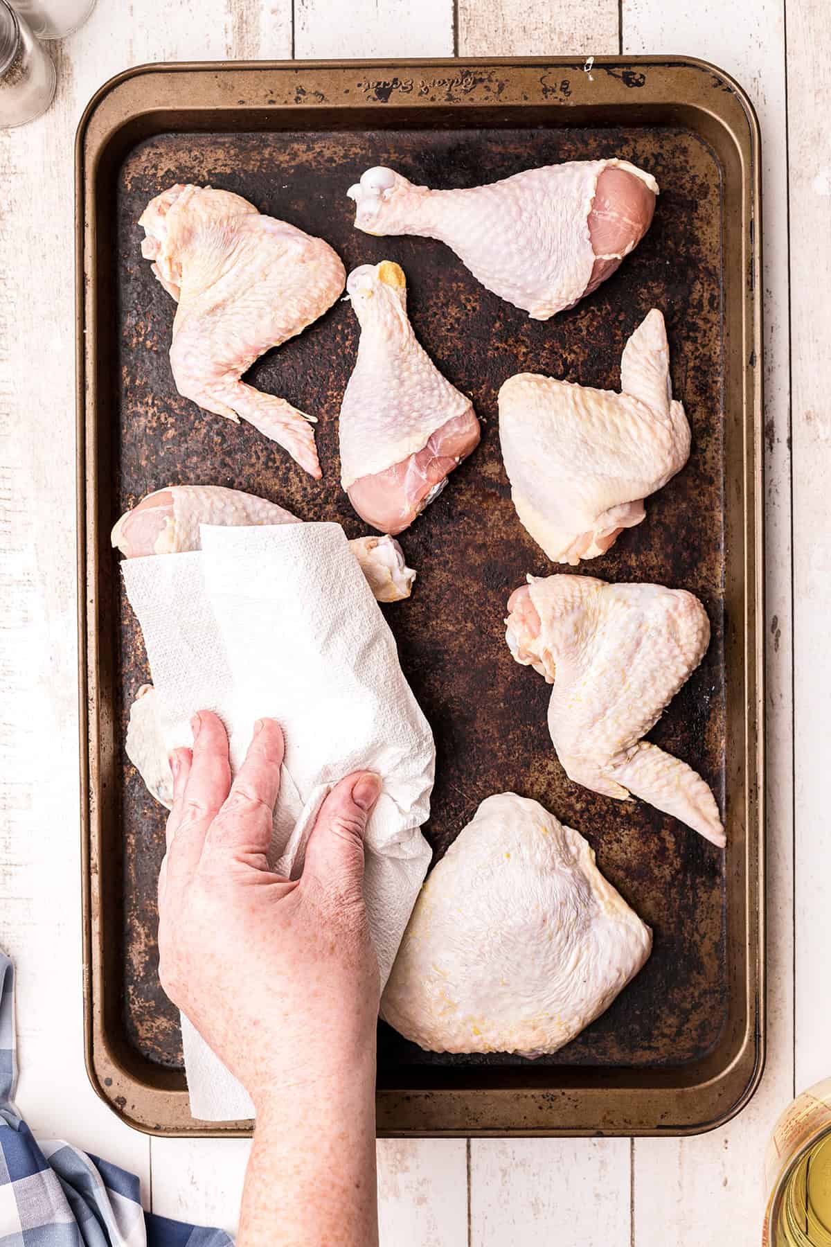 A hand uses a paper towel to pat dry raw chicken pieces on a baking sheet.