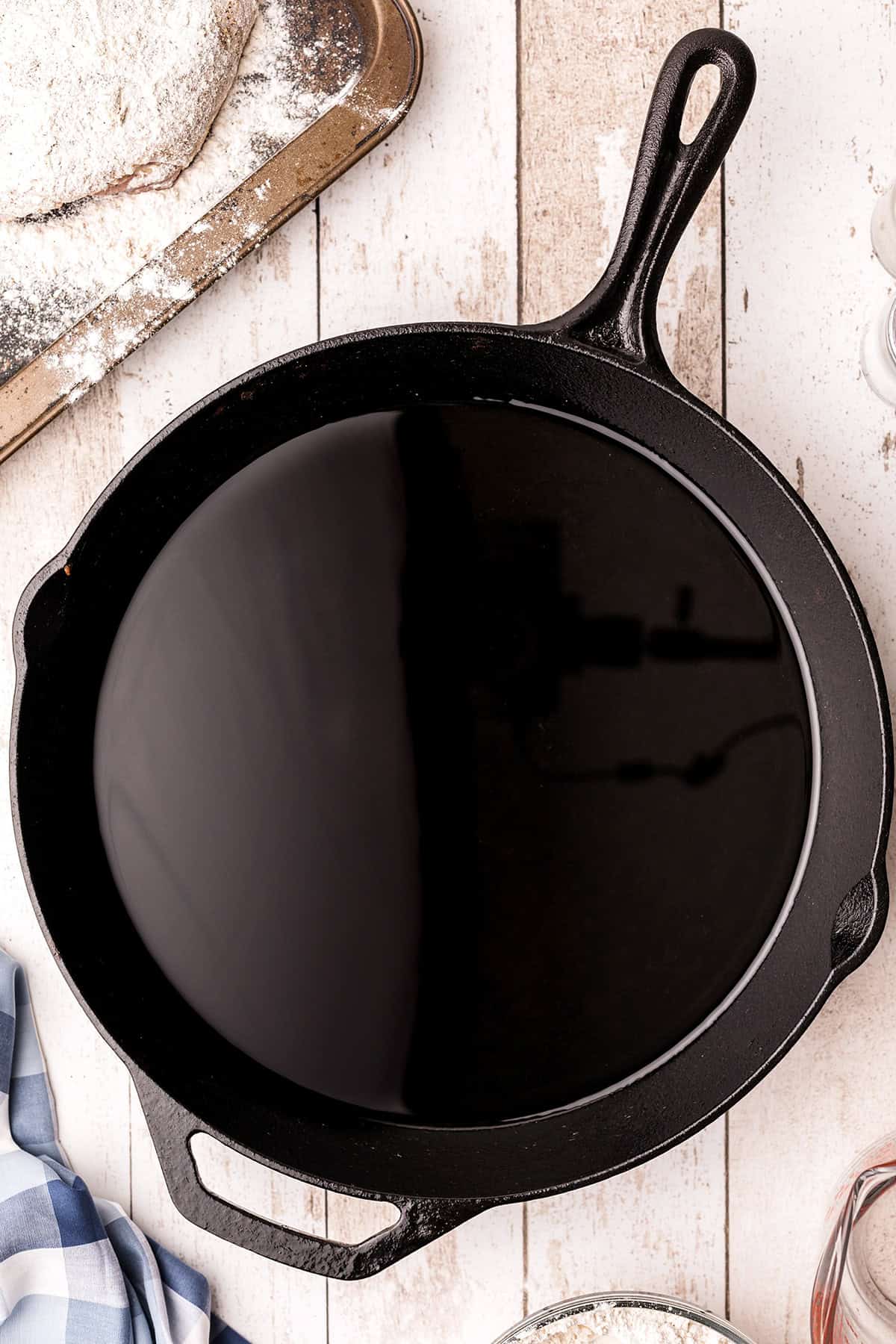 Black cast iron skillet with peanut oil on a white wooden surface next to a baking tray and kitchen towel.