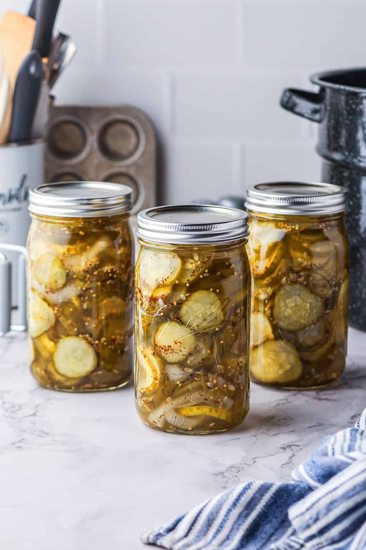 Three jars of homemade pickles on a kitchen counter with utensils and a striped towel nearby.