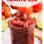 A spoon lifts tomato jam from a glass jar, surrounded by fresh tomatoes and lime slices.