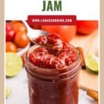 A spoonful of tomato jam above a jar, surrounded by fresh tomatoes, lime halves, and spices.