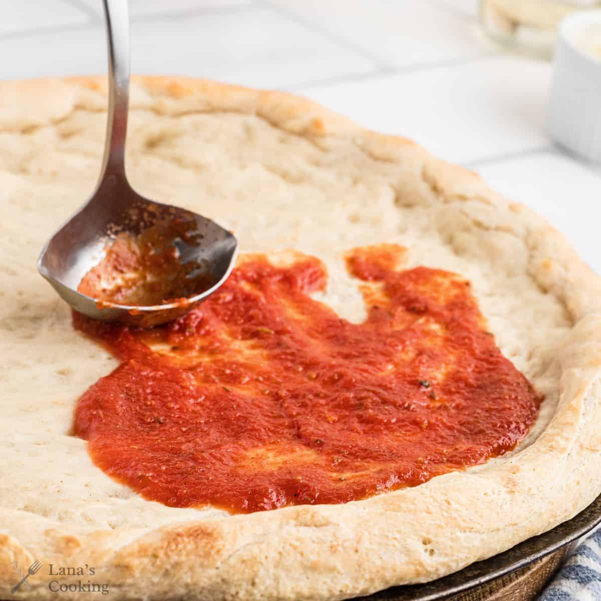 A ladle spreads tomato sauce onto a partially baked pizza crust on a countertop.