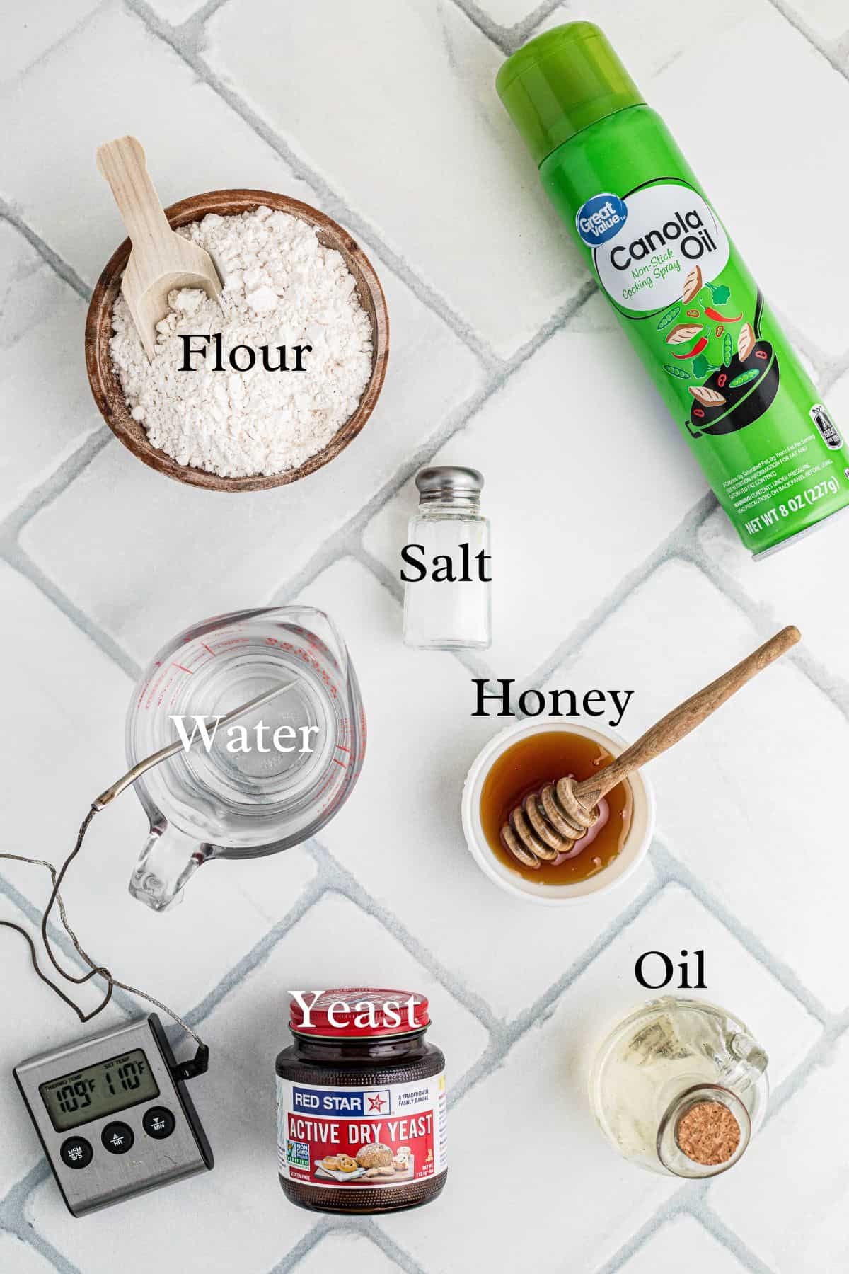A flat lay of bread-making ingredients: a bowl of flour, salt shaker, measuring cup of water, honey jar with dipper, can of canola oil, glass oil jar, dry yeast container, and a thermometer.