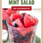 Glass cup filled with watermelon mint salad, including blueberries and grapes, on a green mat.