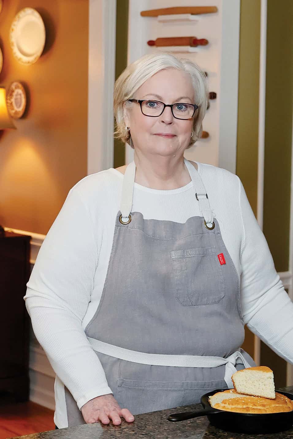 Lana Stuart wearing a gray apron, stands at a kitchen counter with cornbread in a skillet.