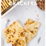 Four spicy, seasoned crackers on a white surface with a dish of crackers and red pepper flakes nearby.