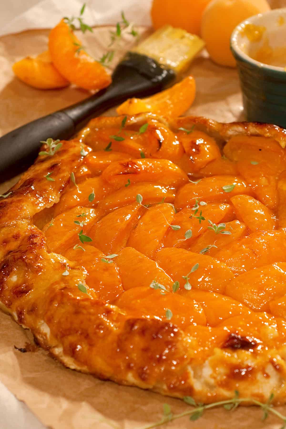 Golden apricot galette garnished with fresh thyme on parchment, with apricot slices and pastry brush nearby.