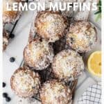 A cooling rack with sugar-dusted blueberry lemon muffins, lemon slices, and scattered blueberries nearby.
