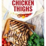 Grilled chicken thighs on a cutting board with herbs, cherry tomatoes, and a plate of salad and tomatoes.