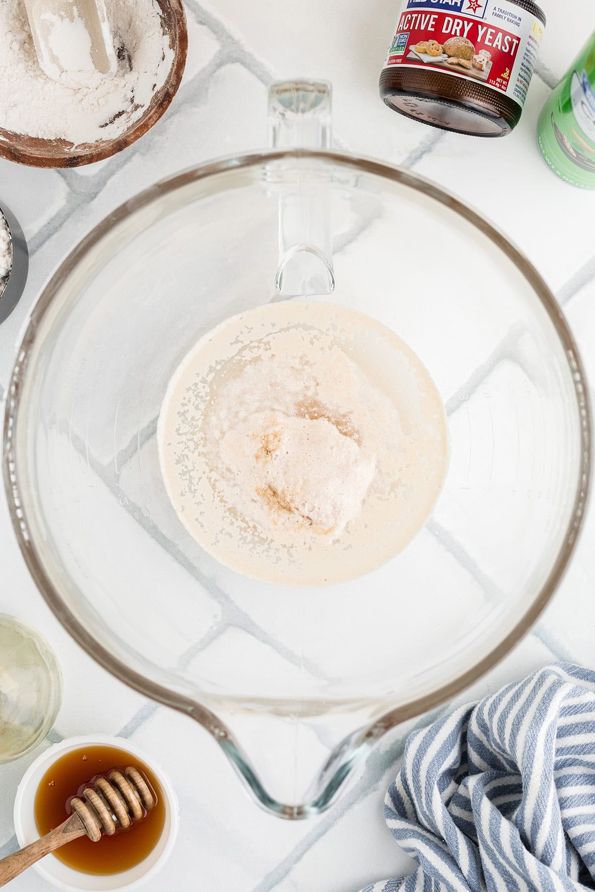 Overhead view of a glass mixing bowl with yeast and flour mixture inside, surrounded by baking ingredients including honey, flour, and a can of active dry yeast.