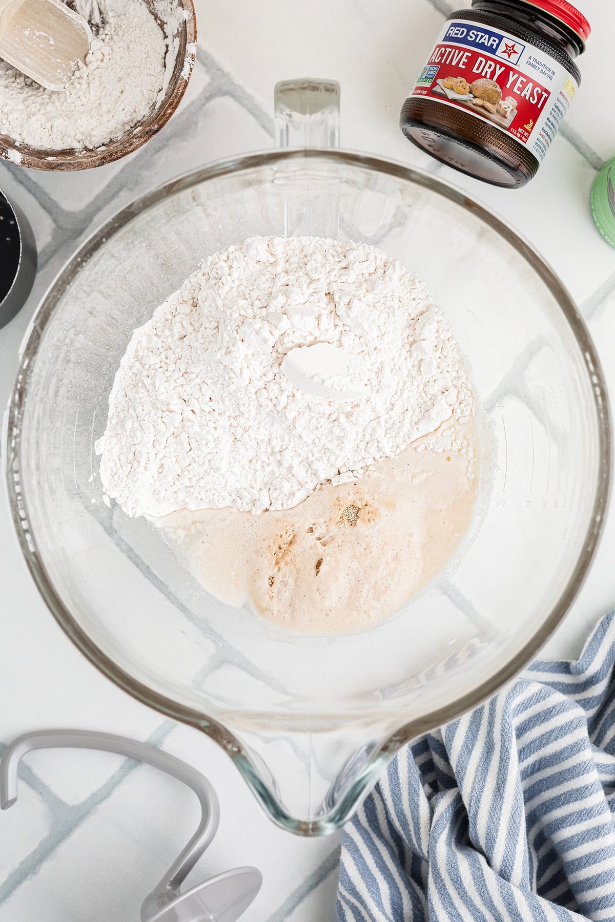 A glass mixing bowl contains flour and yeast mixture on a white countertop, with a jar of active dry yeast, a bowl of flour, and a striped towel nearby.