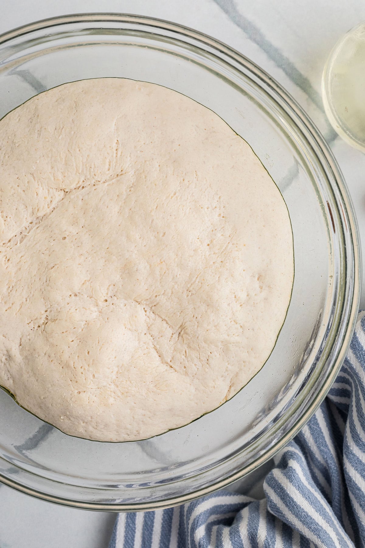 A ball of risen dough in a glass bowl sits on a white surface next to a blue and white striped cloth.