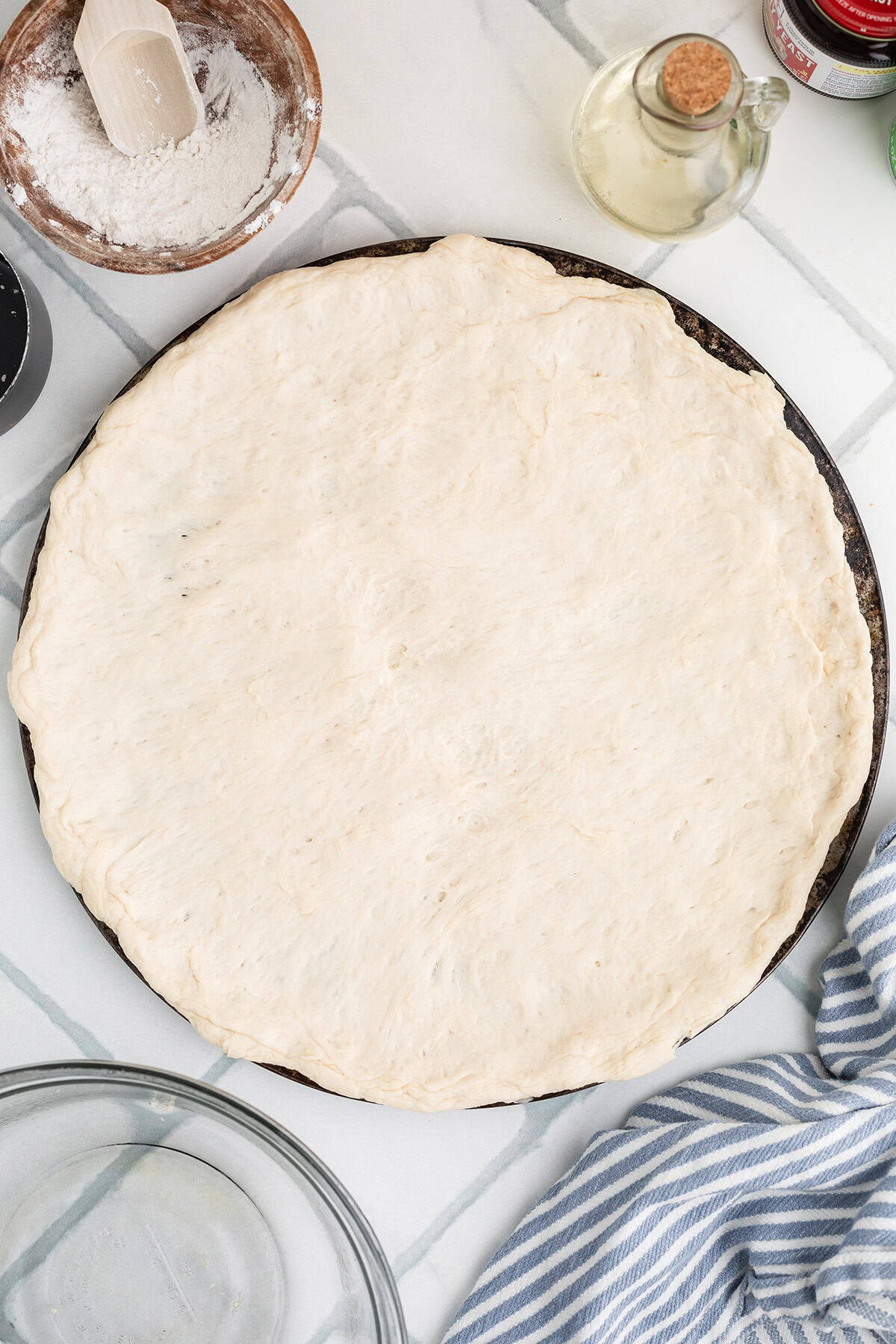 Unbaked pizza dough spread on a round baking tray, surrounded by flour, a rolling pin, a glass bottle of oil, and a striped towel on a light surface.