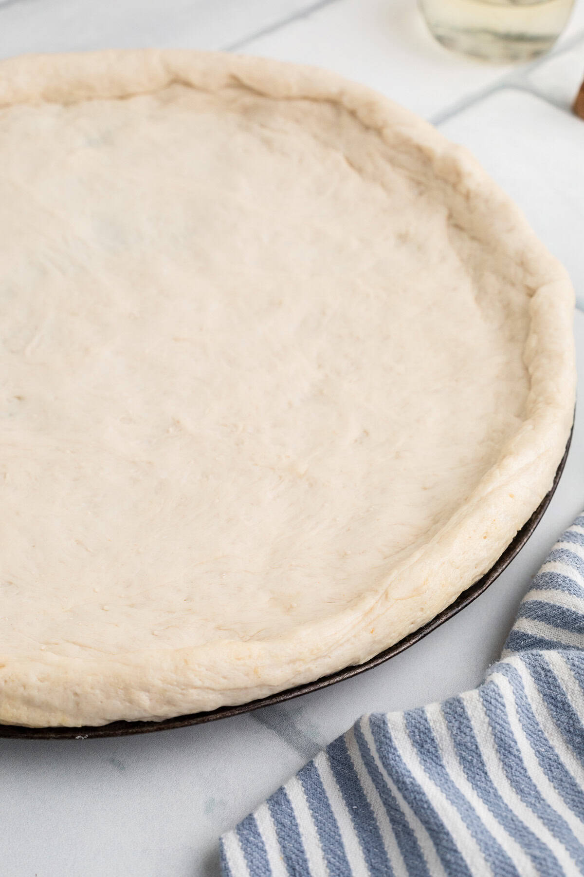 A round, unbaked pizza dough is spread evenly on a pizza pan next to a blue and white striped kitchen towel.