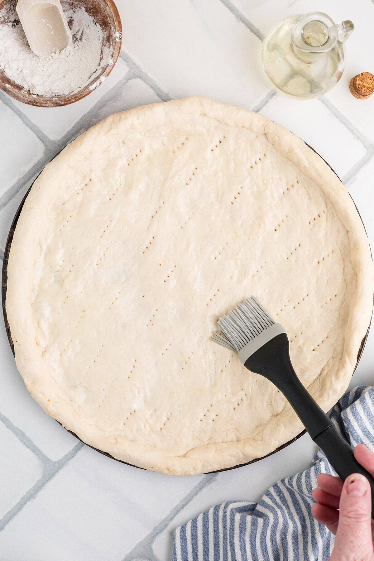 A hand uses a pastry brush to spread oil on a round, uncooked pizza dough that has been docked with fork holes, on a white tiled surface.