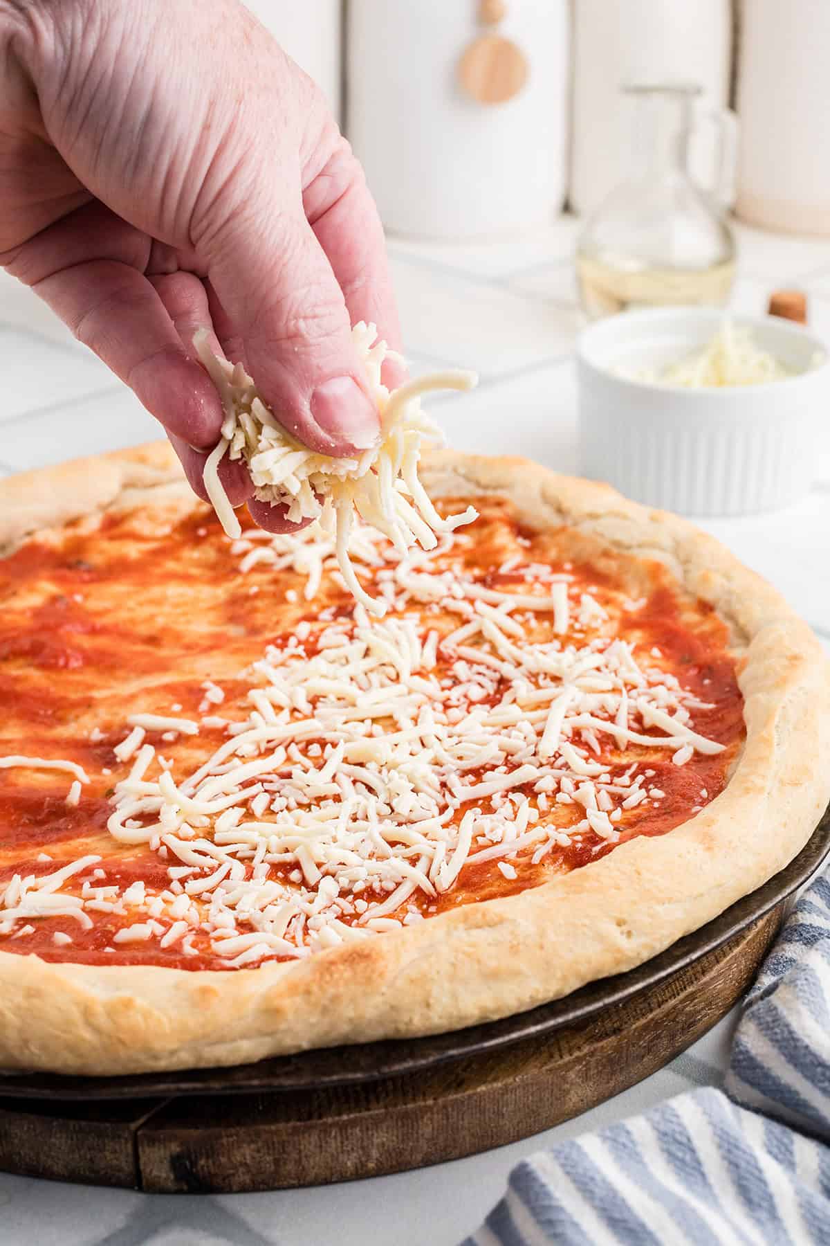A hand sprinkles shredded cheese onto an uncooked pizza with tomato sauce on a wooden pizza board.