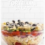 A glass bowl of layered tortellini salad with vegetables, olives, and cheese on a white counter.