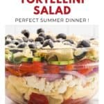 A glass bowl with colorful layered tortellini salad topped with olives, avocado, and grated cheese.