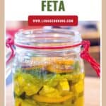 A glass jar filled with marinated feta cubes, herbs, and oil, sealed with a red clasp lid.