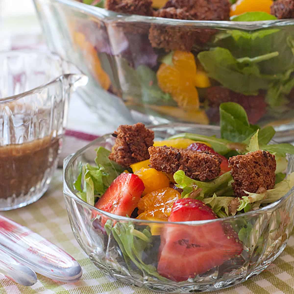 A glass bowl of salad with greens, strawberries, orange slices, and croutons next to a small glass container of dressing.