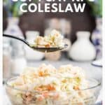 A spoonful of creamy coleslaw is held above a bowl filled with coleslaw, with a blurred kitchen background.