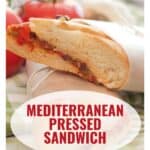 A Mediterranean pressed sandwich filled with vegetables, wrapped in parchment, with tomatoes in the background.