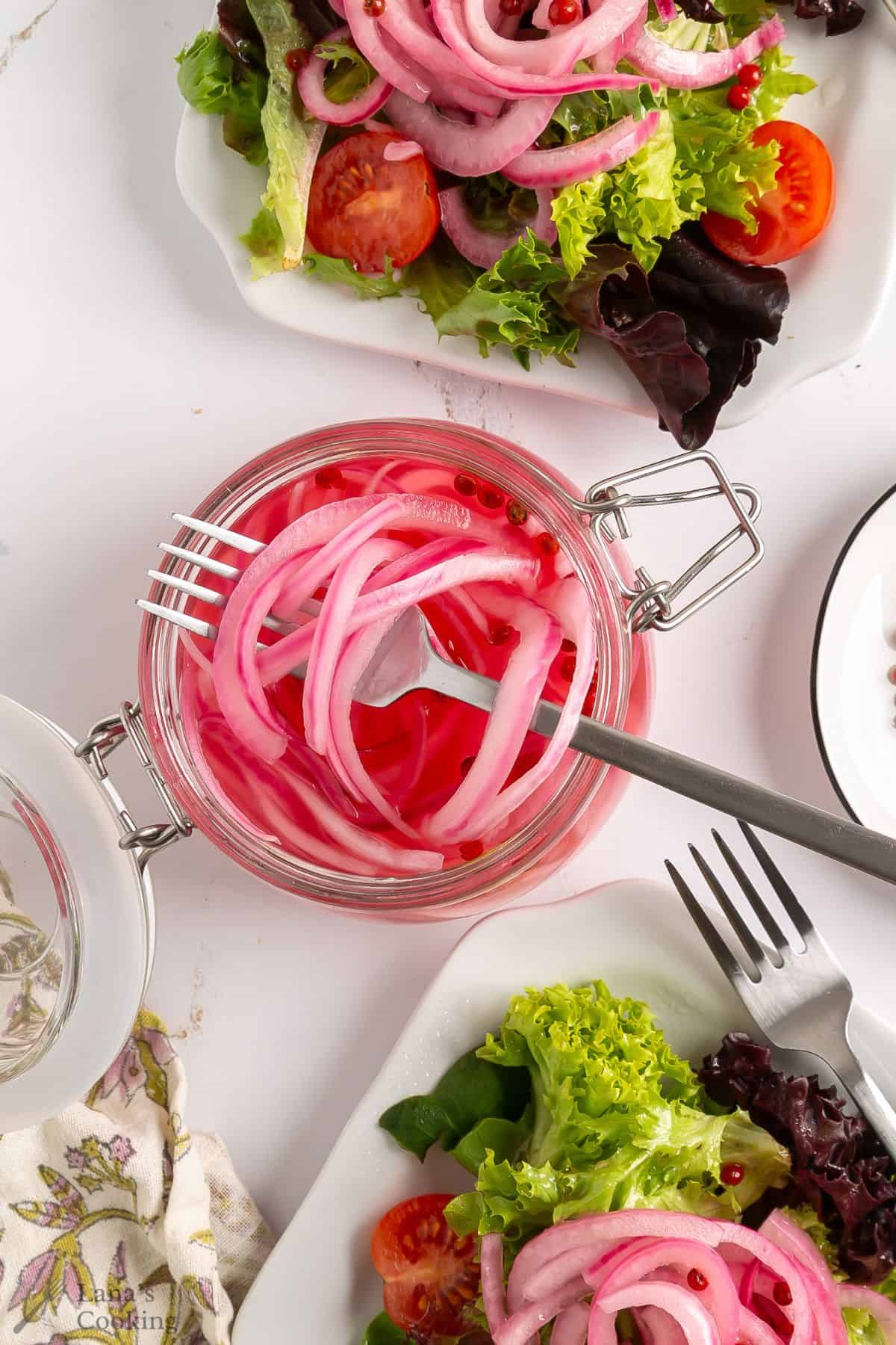 A glass jar filled with pickled red onions with a fork inside, surrounded by plates of fresh green salad with lettuce, tomatoes, and pickled onions, on a white surface with a fork and napkin nearby.